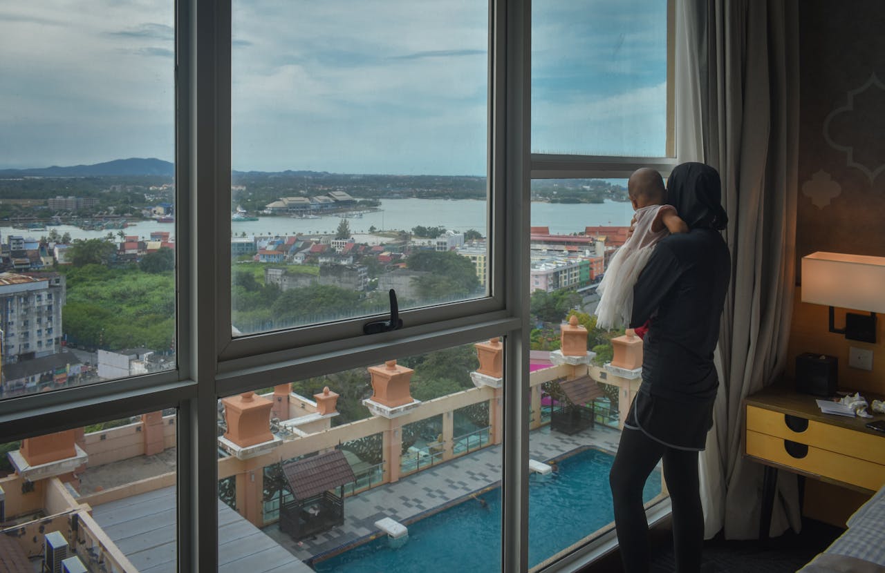 A mother holding her baby gazes out a hotel window at a city view with water in the background.